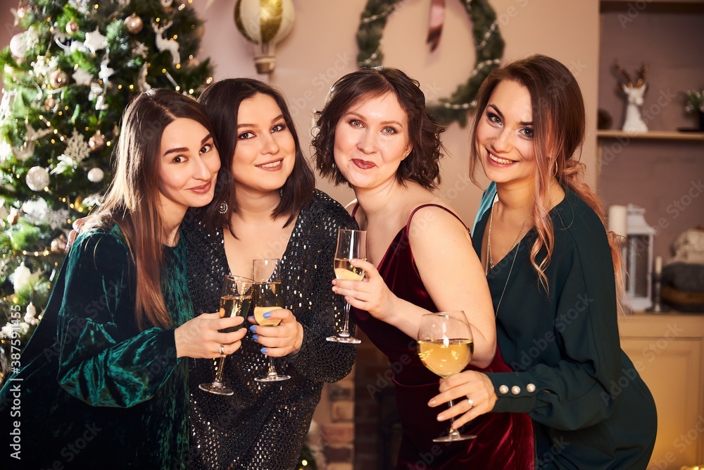 Portrait of four beautiful women in evening dresses who celebrate the new year or Christmas in a beautiful interior with a dressed Christmas tree, garlands and a drink in hand