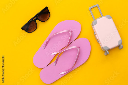 Travel or beach resort flat lay. Mini plastic travel suitcase, sunglasses and flip flops on yellow background. Minimal style. Top view