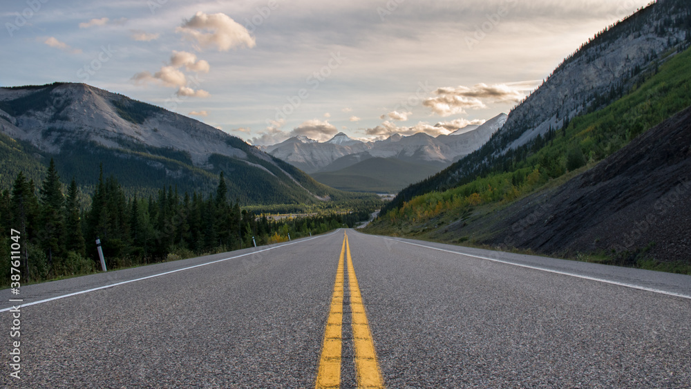 Fototapeta premium Road to the rocky mountains at sunset. Yellow road paint lines wallpaper. Empty deserted street in the countryside rural Alberta valley wallpaper