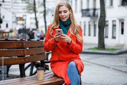 Cheerful young woman tapping and scrolling on smartphone and sitting on bench outside. Stylish attractive female messaging on smartphone. Telephone chatting.