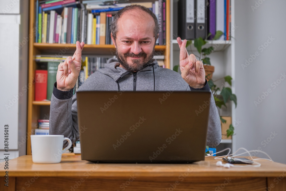 Man working online from home office on computer laptop behind vintage desk holding fingers crossed, listening online meeting with colleagues Man working online from home office on computer laptop behind vintage desk holding fingers crossed, listening online meeting with colleagues