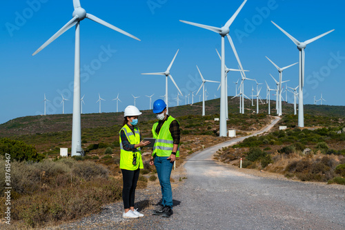 Technical businessman wearing protective face mask working in COVID-19 outbreak after novel coronavirus quarantine and lockdown standing with digital tablet pc on wind power energy station