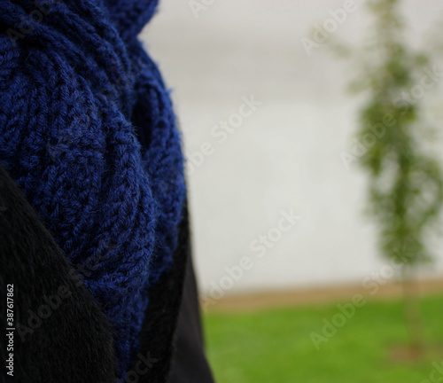 Deep blue knitted woolen scarf on a young man. Black warm coat. Grey wall and green grass background.