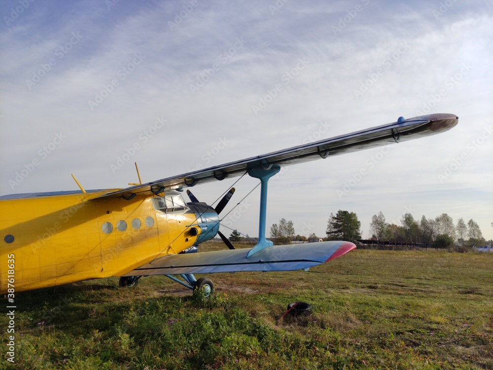 Yellow and blue old biplane plane with a single piston engine and propeller against a blue sky with clouds on the airfield with green grass in summer Yellow and blue old biplane plane with a single piston engine and propeller against a blue sky with clouds on the airfield with green grass in summer