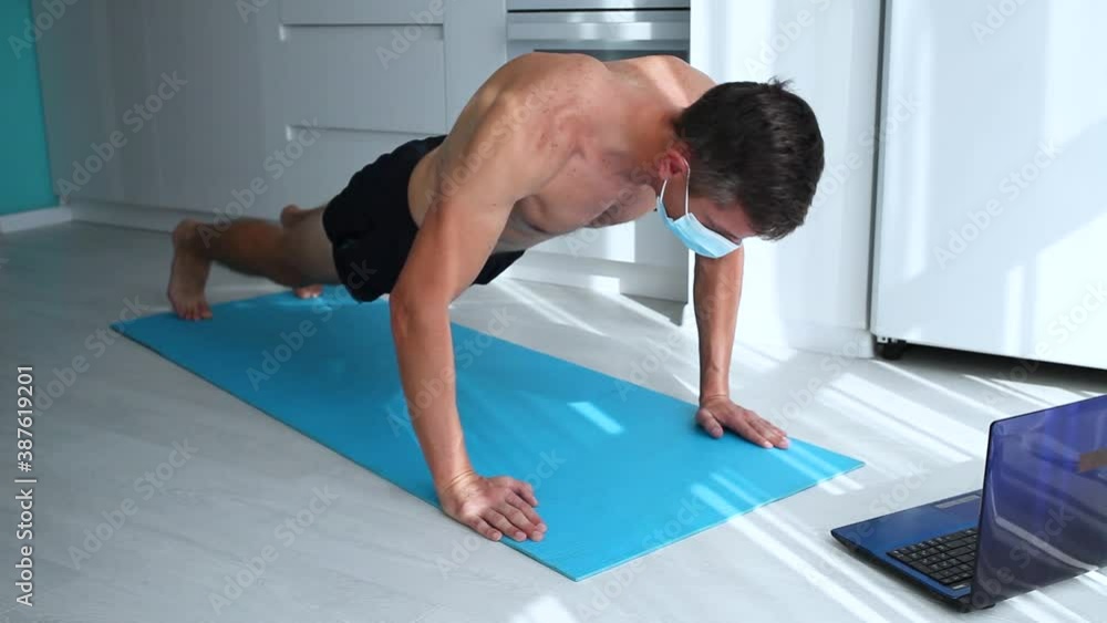 Fit man doing Squeeze-up and watching online lessons on laptop while exercising in the kitchen. Man wearing medical mask Exercise during the COVID-19 Fit man doing Squeeze-up and watching online lessons on laptop while exercising in the kitchen. Man wearing medical mask Exercise during the COVID-19