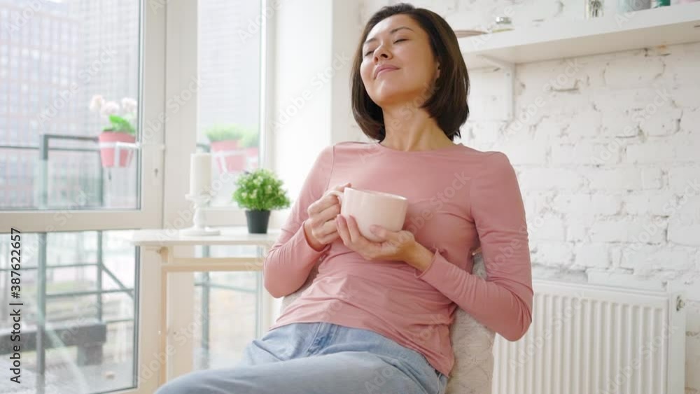 Asian mix race woman enjoying cup of coffee or tea drinking warm beverage at home. Young girl relaxing with morning drink in hands looking through window enjoying and dreaming Asian mix race woman enjoying cup of coffee or tea drinking warm beverage at home. Young girl relaxing with morning drink in hands looking through window enjoying and dreaming