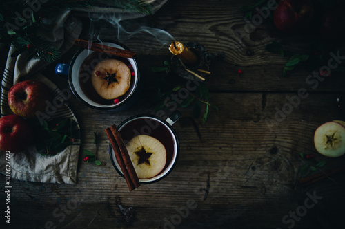 Flat lay with two metal cups with aromatic apple and cranberries tea with cinnamon stick, blown candles, apples and fir branches on old wooden table.