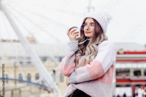 Outdoors lifestyle fashion portrait of stunning blonde young woman. Smiling, drinking hot coffee. Wearing stylish pullover, knitted hat.