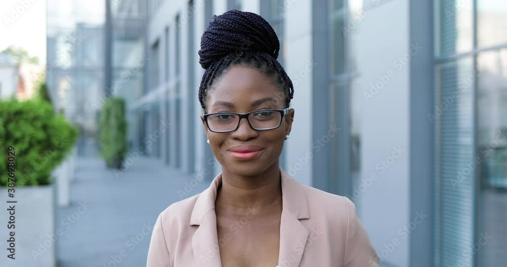 Portrait of young African American beautiful woman in pink jacket and glasses turning face to camera and smiling cheerfully outdoor. Attractive joyful businesswoman at street near business center. Portrait of young African American beautiful woman in pink jacket and glasses turning face to camera and smiling cheerfully outdoor. Attractive joyful businesswoman at street near business center.