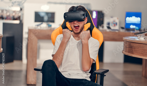 Fotografie Attractive caucasian bearded man trying virtual reality technology while sitting in chair in tech store