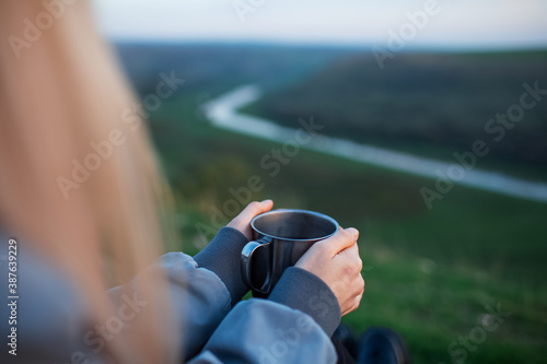 Close-up of female hands holding steel mug with hot tea on background of blurred river.
