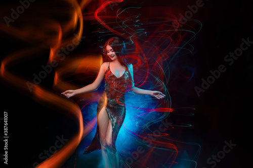 Beautiful girl in motion. Colorful mixed light. Young Attractive happy smiling woman in a shiny evening dress is dancing in night club. Black background, long exposure. Active sexy Lady having fun.