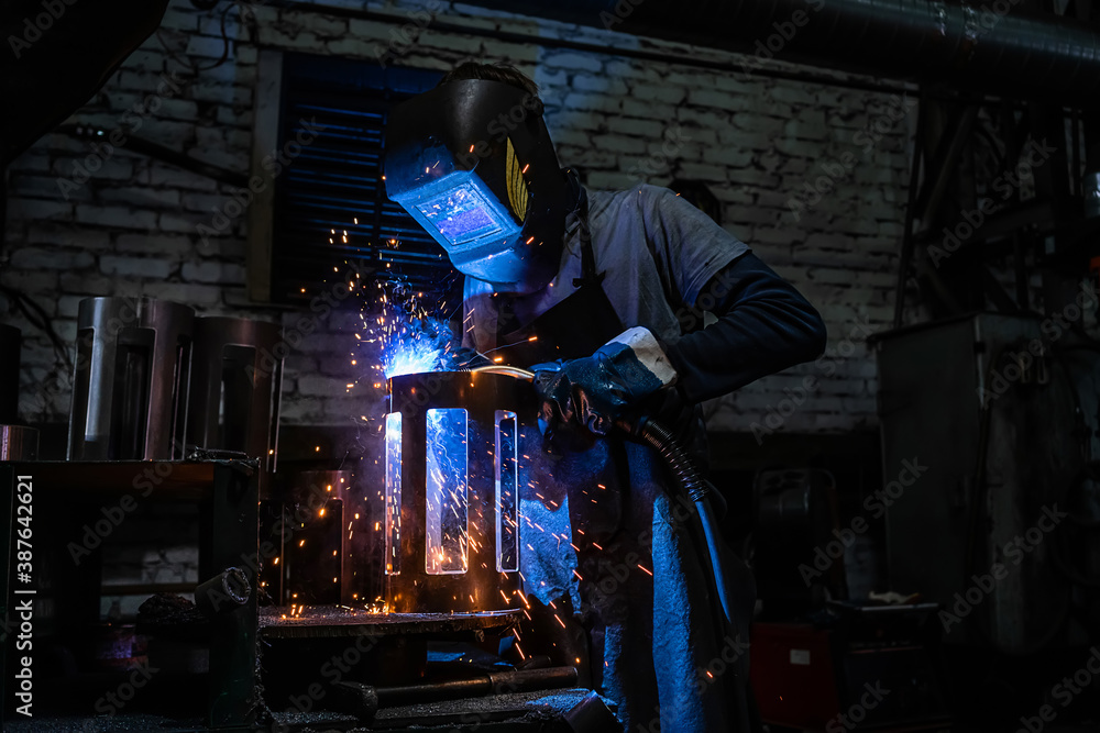 Fototapeta Side view of welder in protective helmet welding metal detail with sparks at factory
