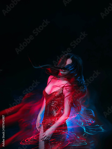 Young beautiful woman in motion. Colorful mixed light. Attractive girl in a shiny evening dress is dancing in night club. Black background, long exposure. Active Lady in sexy silver outfit having fun