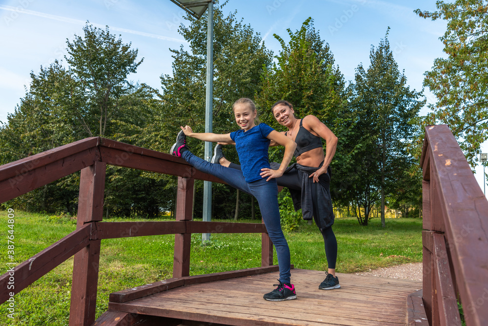 Mother and daughter stretching legs together on top of wooden staircase in public park on sunny summer morning. Family outdoor sports theme. Mother and daughter stretching legs together on top of wooden staircase in public park on sunny summer morning. Family outdoor sports theme.