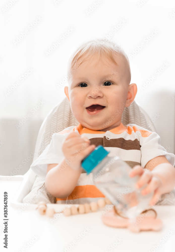 A small surprised blue-eyed child sits at table and looks at the camera. On the table is a bottle with a pacifier and water. Amazement A small surprised blue-eyed child sits at table and looks at the camera. On the table is a bottle with a pacifier and water. Amazement