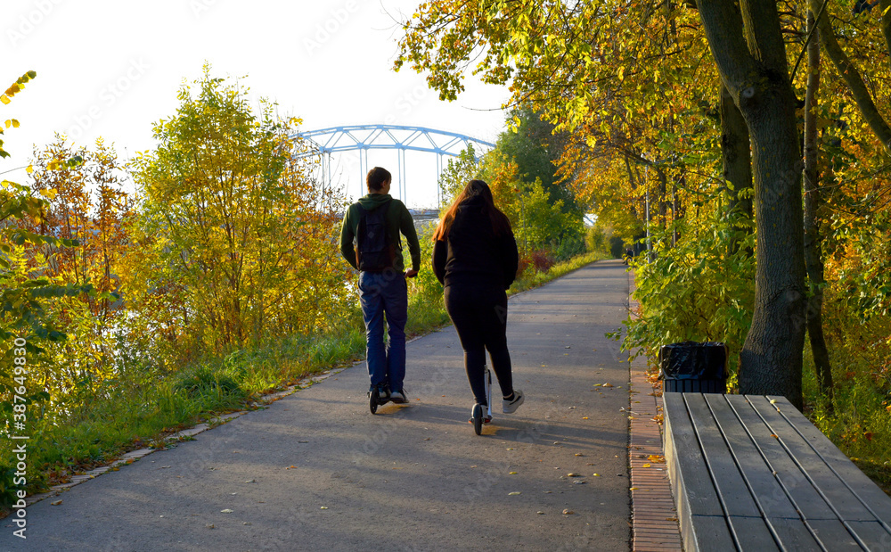 A young man and a girl are walking on scooters on the road of a beautiful Park area. A young man and a girl are walking on scooters on the road of a beautiful Park area.