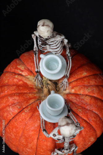 halloween concept - two skeletons drinking tea on pumpkin table against black background flat lay. Vertical image photo