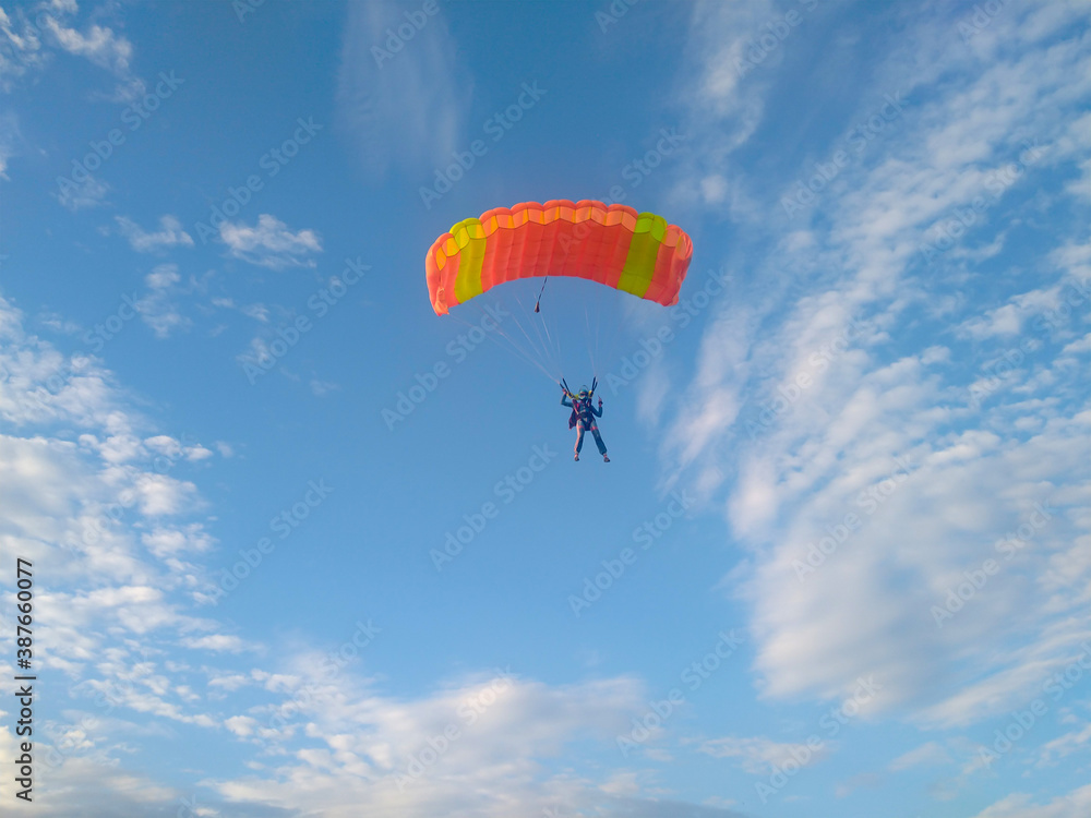 A skydiver with a bright orange sports parachute wing flies before landing in the summer against a blue sky background close up A skydiver with a bright orange sports parachute wing flies before landing in the summer against a blue sky background close up
