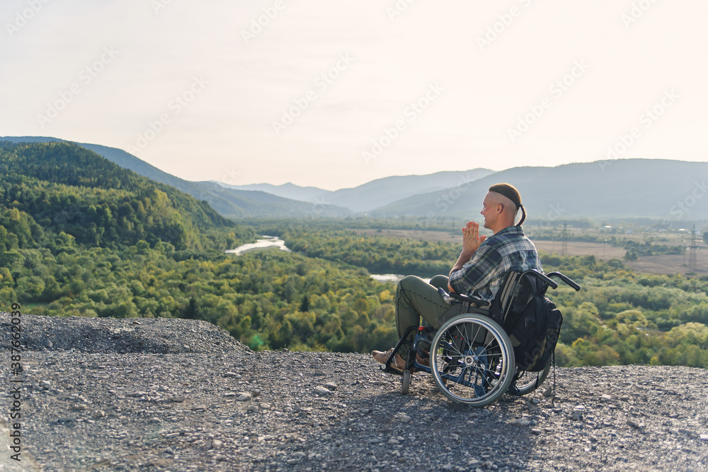 Likable incapacitated young guy sitting alone in wheelchair on the hill on beautiful nature background and praying.