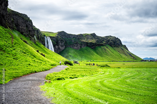 Seljalandsfoss, Iceland waterfall with water cliff in green lush summer rocky landscape and people walking on trail and cloudy dark sky