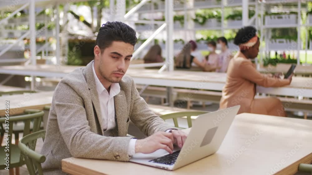 Tracking slowmo of handsome businessman sitting at table in outdoor patio of cafe and typing on laptop, then turning and looking at camera Tracking slowmo of handsome businessman sitting at table in outdoor patio of cafe and typing on laptop, then turning and looking at camera
