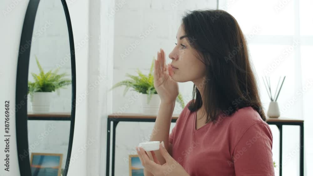 Side view of beautiful young woman applying face cream on skin looking at mirror in bathroom standing alone in light room. People and beauty concept. Side view of beautiful young woman applying face cream on skin looking at mirror in bathroom standing alone in light room. People and beauty concept.