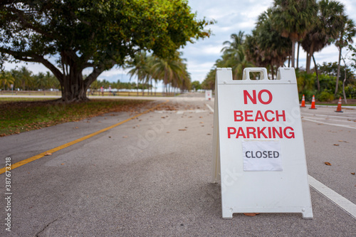Looking across the empty parking lots entrance to a public Key Biscayne beach with leafy trees and locked up park fields in the background, all closed due to the Coronavirus pandemic restrictions