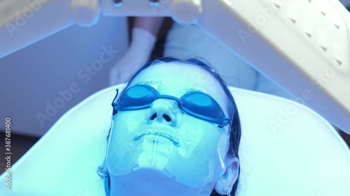 Young woman with collagen hydrogel mask on face having blue LED light facial therapy treatment in beauty salon. Beauty and wellness