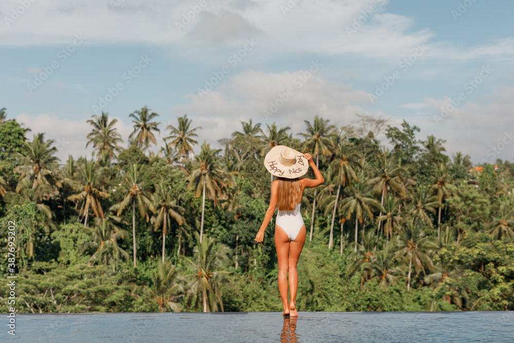Back view young woman in whte swimsuit  and straw hat stands by the pool edge and enjoys the summer with amazing jungle view.  Inspiration travel destination - obrazy, fototapety, plakaty 