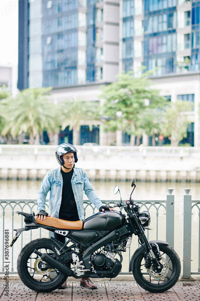 Young serious Asian man in helmet standing on bridge next to his motorcycle Young serious Asian man in helmet standing on bridge next to his motorcycle