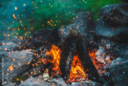 Vivid smoldered firewoods burned in fire close-up. Atmospheric background with orange flame of campfire. Full frame image of bonfire. Warm whirlwind of glowing embers and ashes in air. Sparks in bokeh