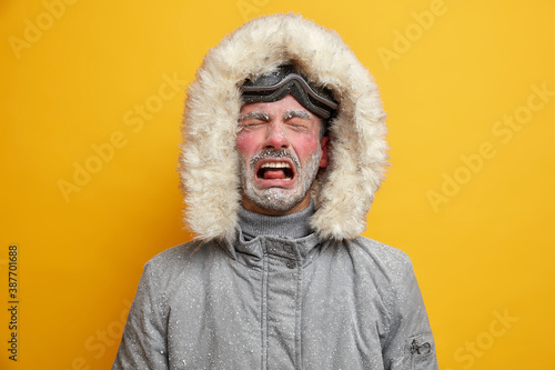 Dejected man cries and feels unhappy dressed in winter clothes feels very cold after snowboarding has red face covered with hoarfrost wears ski goggles isolated over yellow wall. Recreation concept