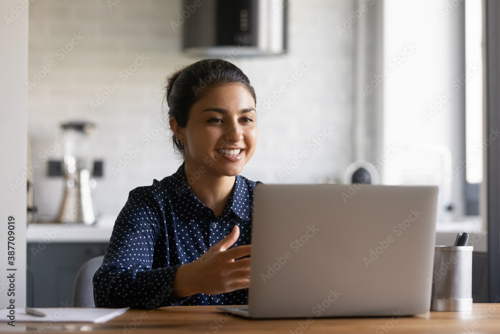 Smiling millennial Indian female look at laptop screen speak talk on video call on gadget. Happy young ethnic woman working on modern computer at home office. Online communication, technology concept. Smiling millennial Indian female look at laptop screen speak talk on video call on gadget. Happy young ethnic woman working on modern computer at home office. Online communication, technology concept.