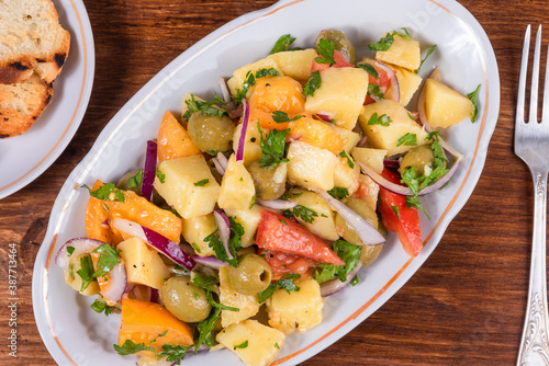 Traditional Italian Calabrian potato salad - oval plate with boiled potatoes, tomatoes, onions and olives on a plain table close-up photo