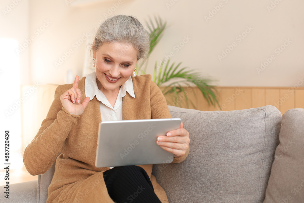 Cheerful middle aged woman sitting on sofa, using computer tablet apps, looking at screen,reading good news in social network, shopping or chatting online. Cheerful middle aged woman sitting on sofa, using computer tablet apps, looking at screen,reading good news in social network, shopping or chatting online.