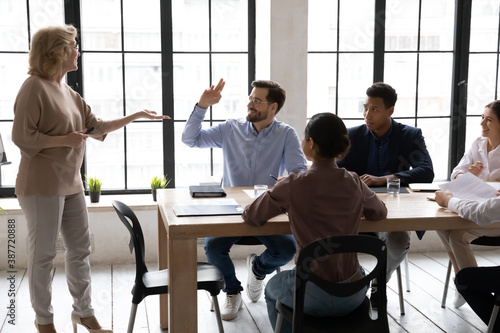 Smiling Caucasian male employee raise hand ask answer question at meeting with middle-aged businesswoman or CEO. Confident motivated man worker participate in team discussion at briefing.