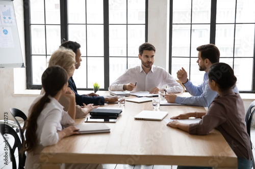 Young Caucasian businessman head lead meeting with multiracial diverse employees in office. Multiethnic businesspeople talk brainstorm discuss business ideas at team briefing. Teamwork concept.