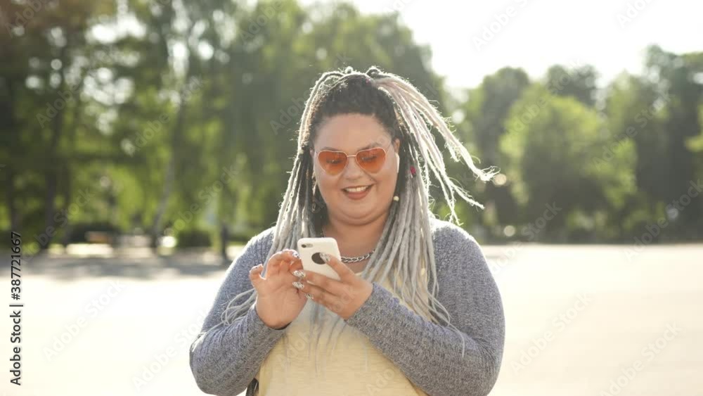 Smiling happy woman with dreads texting message standing at city street during summertime. Stout girl or mobile user looking at mobile screen surfing in internet. Lifestyle and social media concept Smiling happy woman with dreads texting message standing at city street during summertime. Stout girl or mobile user looking at mobile screen surfing in internet. Lifestyle and social media concept