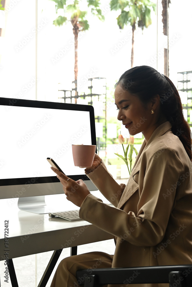 A young Asian businesswoman working on computer and reading incoming notification on smartphone at office. A young Asian businesswoman working on computer and reading incoming notification on smartphone at office.