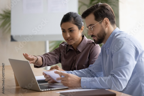 Diverse multiracial colleagues sit at desk look at laptop screen discuss company financial project together. Serious multiethnic coworkers work collaborate brainstorm using computer at office meeting.