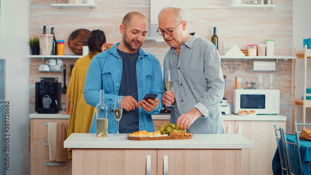Young man showing his father how to use modern smartphone sitting in the kitchen and drinking a glass of white wine before family dinner. New lifestyle, using technology while women preparing the Young man showing his father how to use modern smartphone sitting in the kitchen and drinking a glass of white wine before family dinner. New lifestyle, using technology while women preparing the