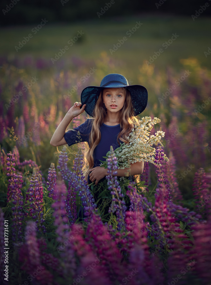 Happy young girl in blue dress and hat with violet flowers in the summer blooming field watcing at the camera