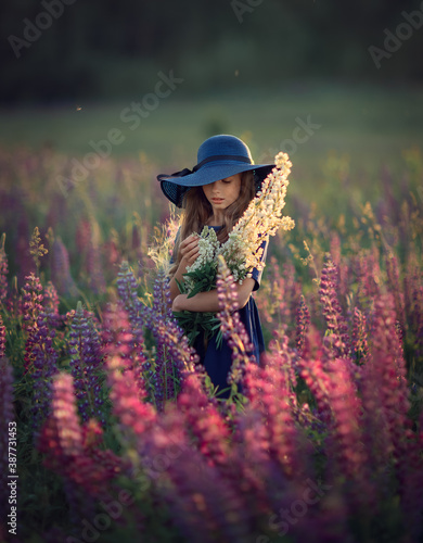 Happy young girl in blue dress and hat with violet flowers in the summer blooming field