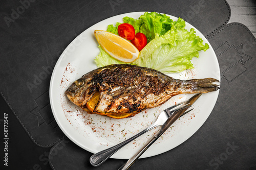 Grilled dorado fish stuffed with orange, garnished with lettuce, lemon and cherry tomato, knife and fork, top view