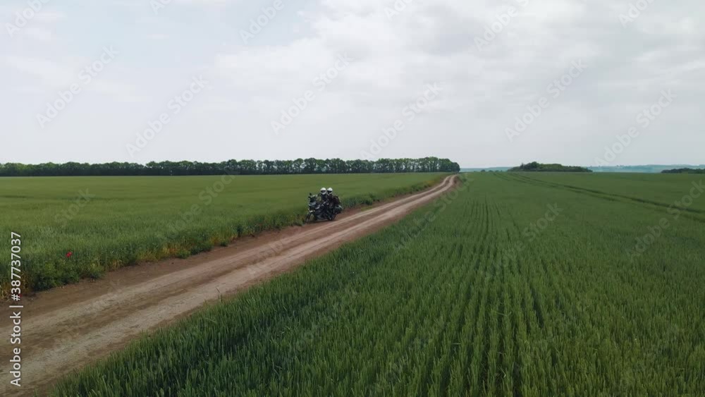 A couple of motorcyclists ride a touristic motorcycle on a dirt road. Green fields beyond the horizon. Summer day. The concept of outdoor recreation. Weekends. Vacation and extreme hobbies. Aerial vie A couple of motorcyclists ride a touristic motorcycle on a dirt road. Green fields beyond the horizon. Summer day. The concept of outdoor recreation. Weekends. Vacation and extreme hobbies. Aerial vie
