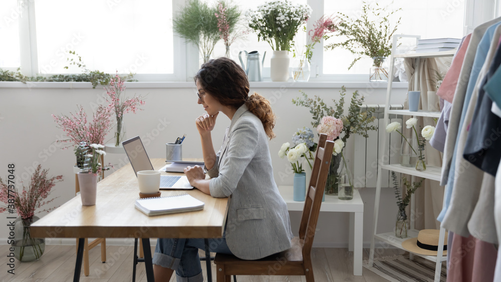 Millennial Caucasian female florist sit at home office or atelier look at  laptop screen working distant. Young woman designer or decorator use  computer consult client customer online on gadget. Stock Photo