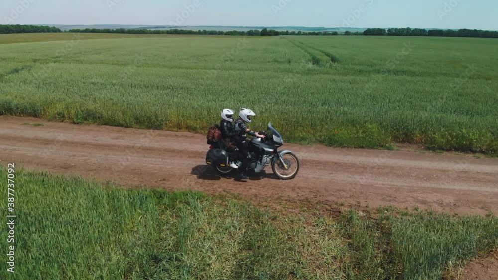 A couple of motorcyclists ride a touristic motorcycle on a dirt road. Green fields beyond the horizon. Summer day. The concept of outdoor recreation. Weekends. Vacation and extreme hobbies. Aerial vie A couple of motorcyclists ride a touristic motorcycle on a dirt road. Green fields beyond the horizon. Summer day. The concept of outdoor recreation. Weekends. Vacation and extreme hobbies. Aerial vie