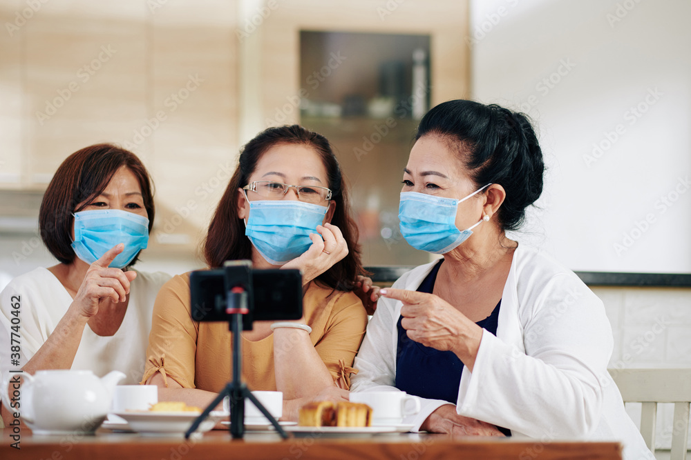 Cheerful Asian women in medical masks filming podcast with themselves discussing news at kitchen table