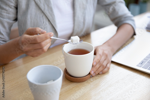 Crop close up of woman sit at desk put cube of white sugar drinking warm hot tea at workplace. Female employee sweeten beverage, enjoy leisure break time at work. Hygge, dieting concept.